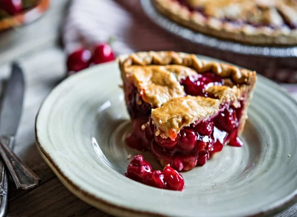 closeup of slice of cherry pie on plate with whole pie in background